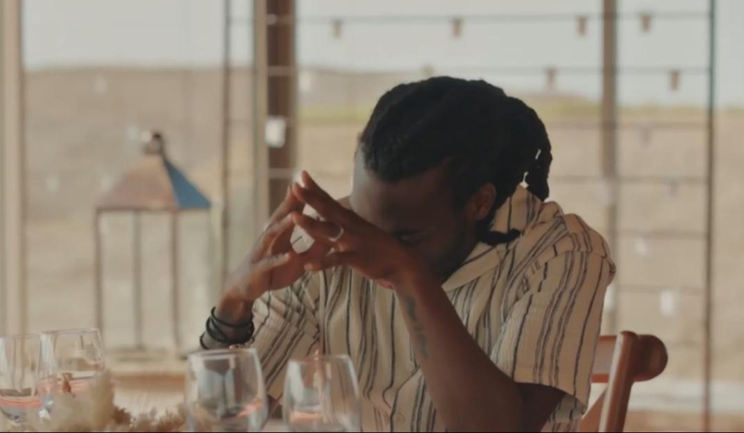 A man wearing a striped shirt sits at a table with his hands clasped over his face.