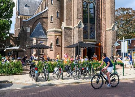 Patrons seated at outdoor tables at a cafe next to a brick church; a cyclist rides by.