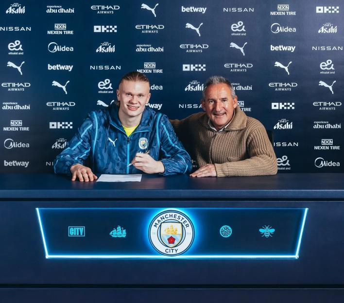 Erling Haaland and Txiki Begiristain sitting at a table with Manchester City logos, signing a contract.