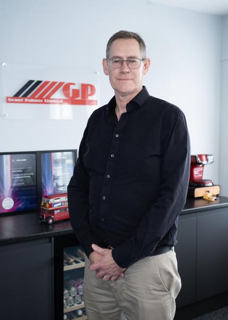 A man in glasses, a black shirt, and khaki pants stands in an office with the "Grant Palmer Limited" logo behind him.