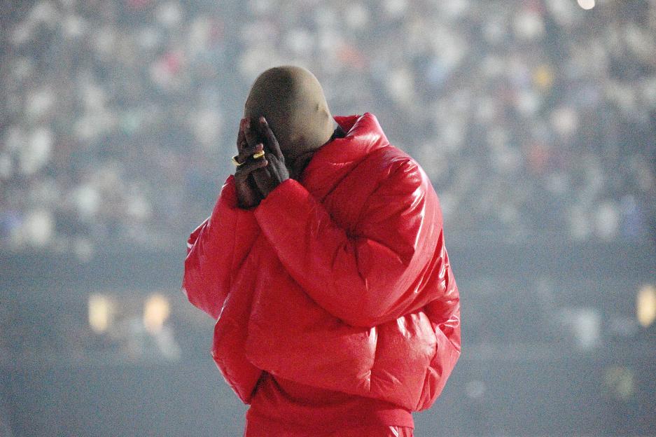 Kanye West in a red puffer jacket with a face mask, hands clasped, at a listening event.