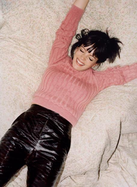 Lily Allen laying on a floral patterned bedspread, laughing with her arms stretched above her head, wearing a pink sweater and dark pants.