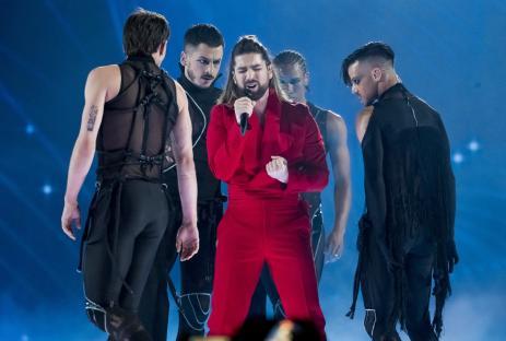 A singer in a red suit performs on stage with backup dancers.