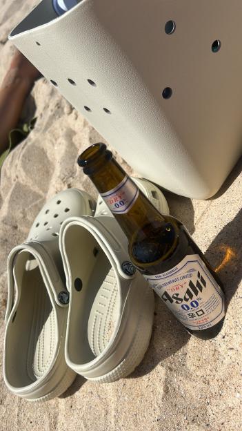 A non-alcoholic Asahi beer bottle, Crocs, and a bag are on the sand.