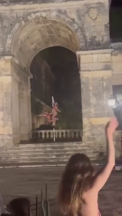 Two pole dancers performing on a pole in front of a stone archway.