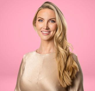 Portrait of a smiling woman with long blonde hair wearing a gold top against a pink background.