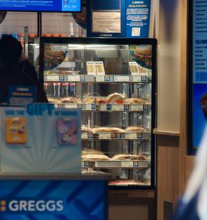 Interior of a Greggs store showing refrigerated sandwiches and drinks moved behind the counter to deter shoplifting.