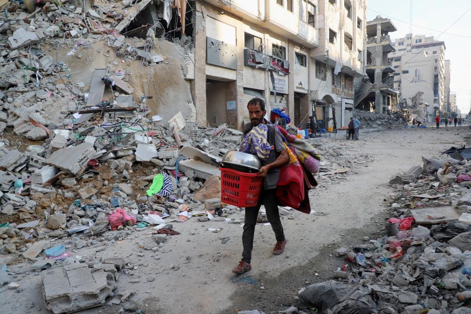 A Palestinian man carries belongings through a destroyed city street.