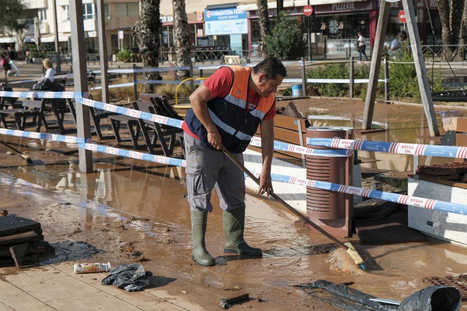 A man in a safety vest and rubber boots uses a broom to clear floodwater from a street in Ibiza, Spain.