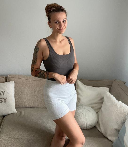 A woman with tattoos on her arms smiles at the camera in a gray top and light gray shorts.