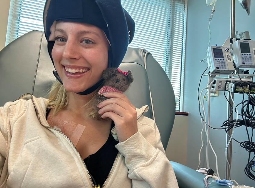 A smiling woman in a hospital room during cancer treatment holds a small stuffed animal.