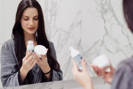 Woman in bathrobe examining skincare products in bathroom mirror.
