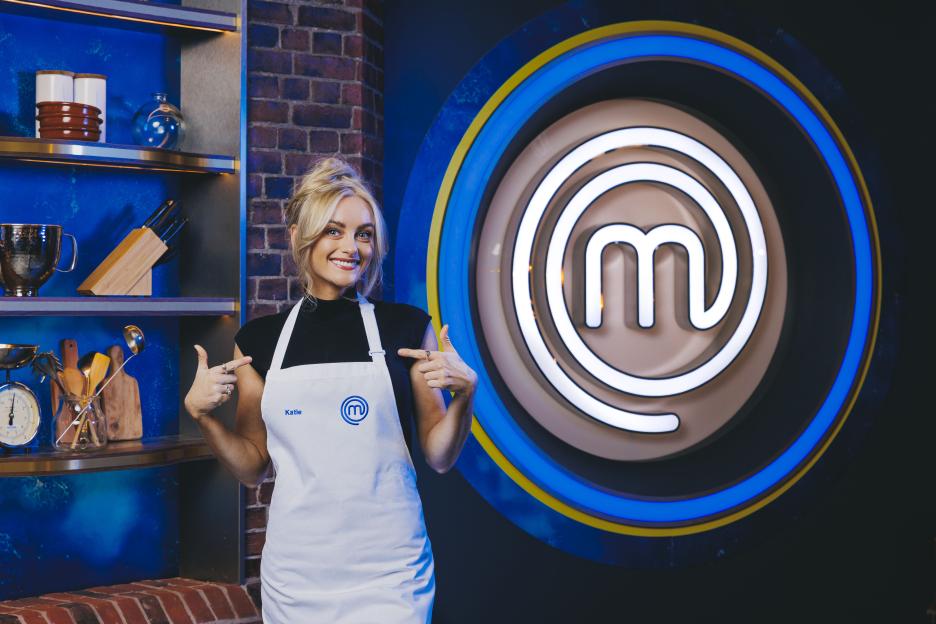 Katie, a contestant on Celebrity MasterChef S20, pointing to her apron with the MasterChef logo.
