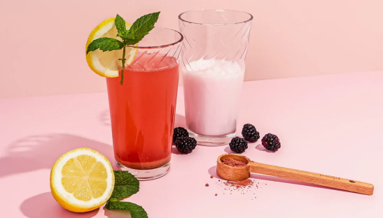 Two glasses of pink and red drinks with lemon and mint garnish, blackberries, and a wooden scoop of powder.