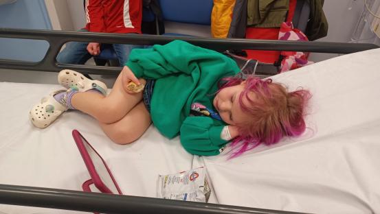 Young girl with pink hair lying on a hospital bed, eating a snack.