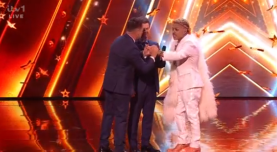 A performer in a white suit shakes hands with a judge on a televised talent show stage.