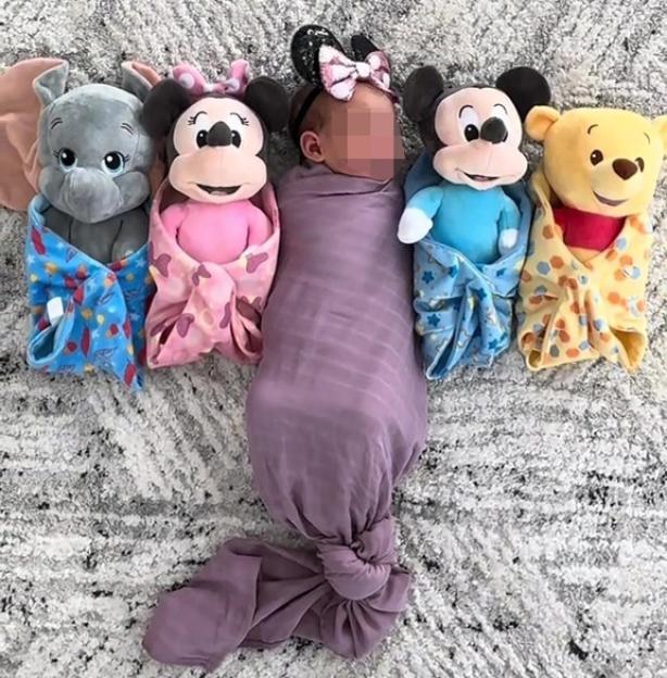 Newborn baby, Disney Mae Jones, swaddled in purple and surrounded by four Disney plush toys.
