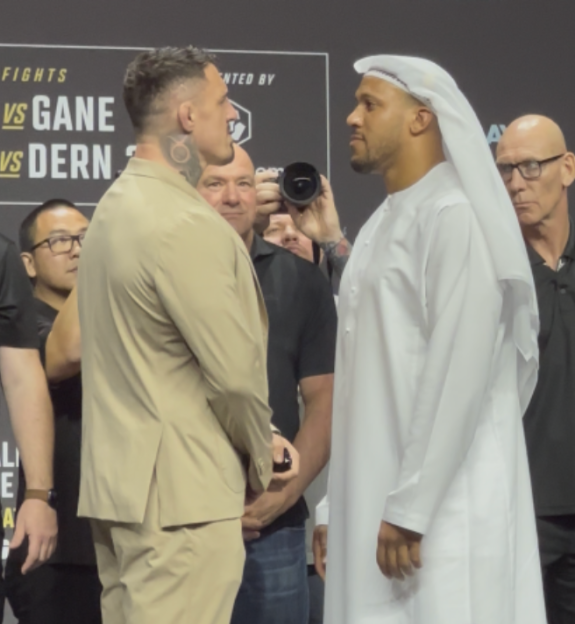 Two male fighters facing each other at a weigh-in, one dressed in a tan suit, the other in a white thawb.