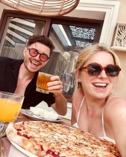 A man and woman smiling at the camera while enjoying pizza and drinks outdoors.