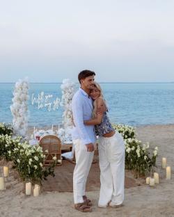 Luca Bish and Grace Jackson embracing on a beach with a proposal sign.