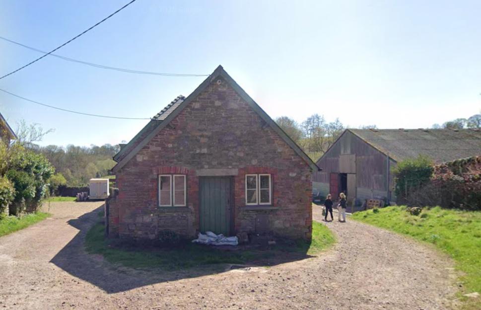 A stone barn with a green door and two windows sits on the left of a dirt path. To the right is another large barn. Two women are walking away from the camera on the path towards the larger barn.