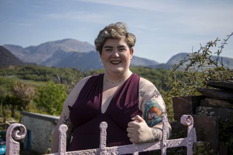 A woman stands by a gate, smiling at the camera. Mountains are visible in the background.
