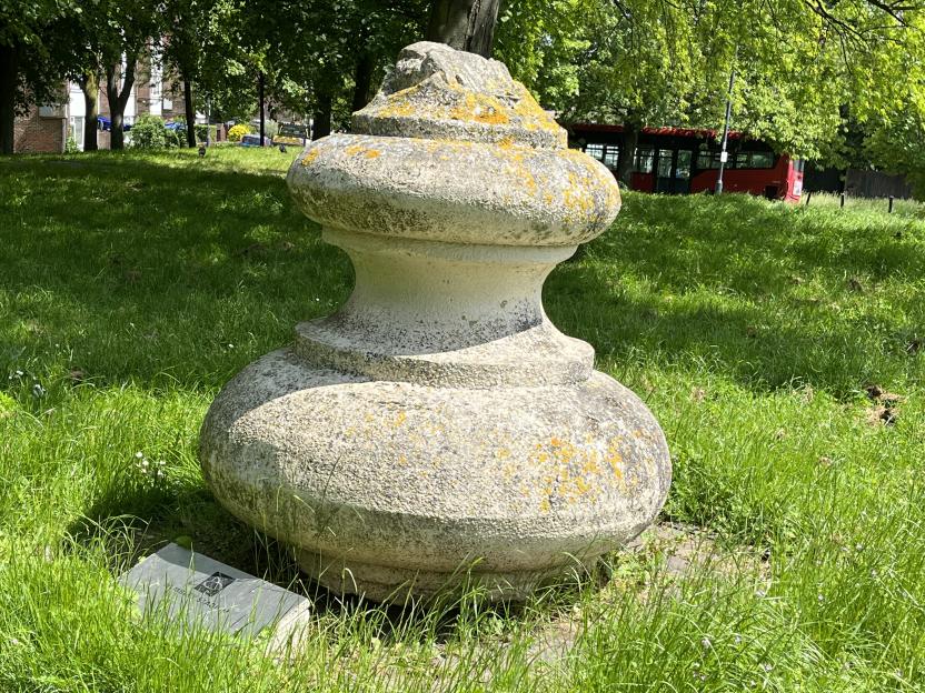 A large, weathered stone finial with lichen on it, resting on a grassy area, with a red double-decker bus visible in the background.