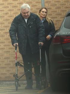 A man using a walker walks with a woman beside a car.