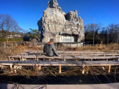 Person sitting on a bench in front of an abandoned Mount Rushmore-like structure at a derelict theme park.