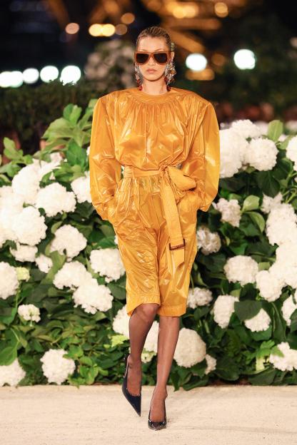 Bella Hadid walks the runway in an orange shiny jumpsuit with matching belt, large sunglasses, and black heels.