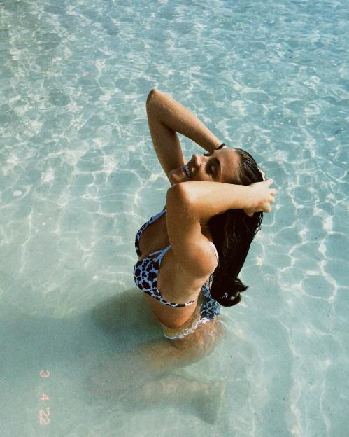 Woman in cow print bikini sitting in shallow, clear water.
