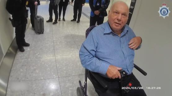 An elderly man in a wheelchair being escorted by police.