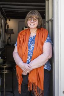 A woman stands in a doorway, wearing an orange shawl and a floral shirt.
