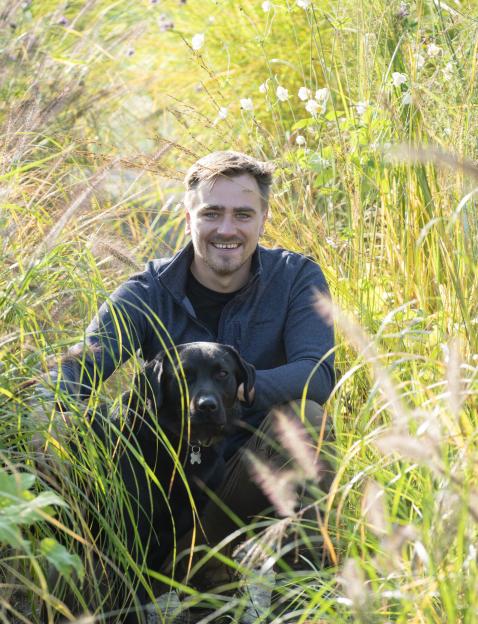 Man and black dog sitting in tall grass.