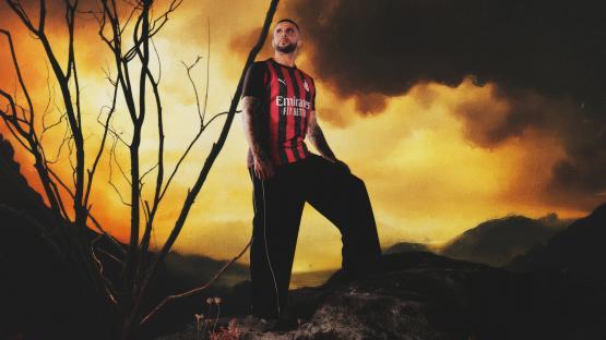 A man in the AC Milan 2025/26 jersey stands on a rock against a dramatic sunset.
