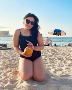 Woman in a swimsuit kneeling on a beach, drinking from a coconut.
