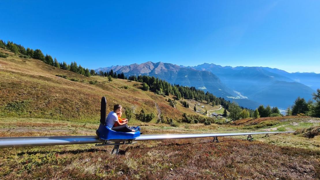 People in a toboggan ride on a track on a mountain slope with trees and distant mountains.