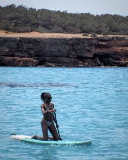Woman in black swimsuit sitting on a boat, with sailboats in the background.