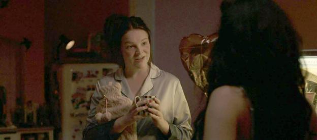 Woman in pajamas holding a mug and a stuffed animal, talking to another woman.