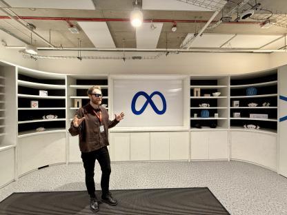 Man in glasses gesturing in front of a Meta logo displayed on a wall with shelves.