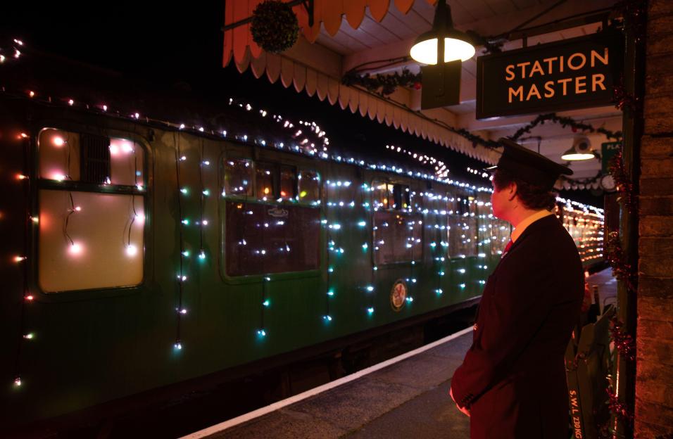 A guard in a hat looks at a green train adorned with colorful LED lights at a station at night.