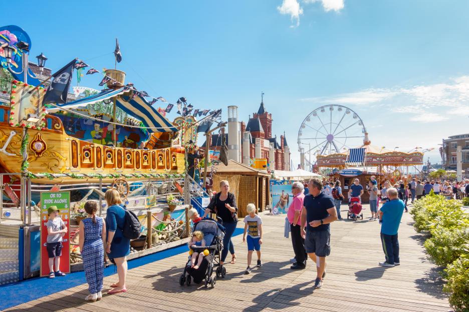 People enjoying a sunny day at a Cardiff Bay fair with rides and a Ferris wheel.
