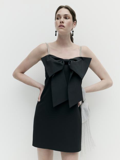 Woman models a black mini-dress with a large bow detail and crystal-embellished straps for the M&S Autumn Winter 2025 collection.