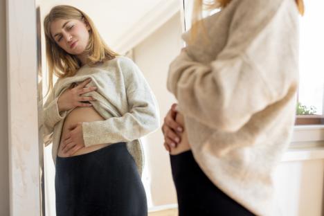 Woman looking in mirror, holding her abdomen in pain.