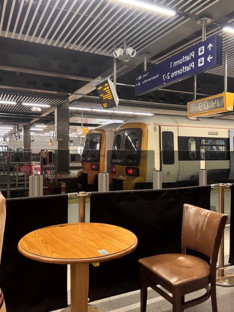 A wooden table and brown chair in the foreground of a train station platform with two trains stopped at the platform.