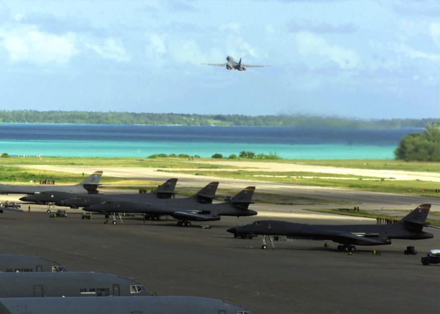 Photo of B-1 bombers on a runway with a B-1 taking off in the background, overlooking a tropical atoll.