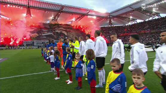 Soccer players and children on the field during a game with red flares going off in the stands.