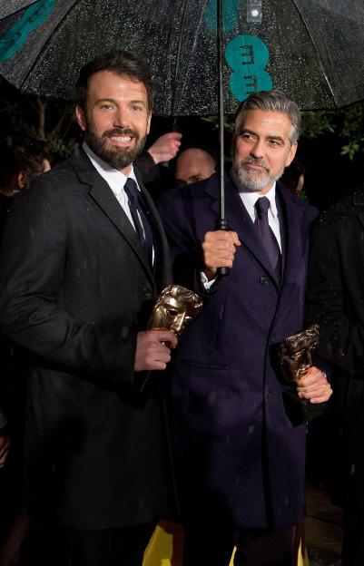 Ben Affleck and George Clooney holding BAFTA awards under an umbrella.