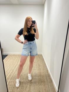 Woman in light denim shorts and black top taking a mirror selfie.