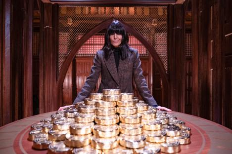 Claudia Winkleman on the set of The Traitors, standing behind a large pile of gold coins.